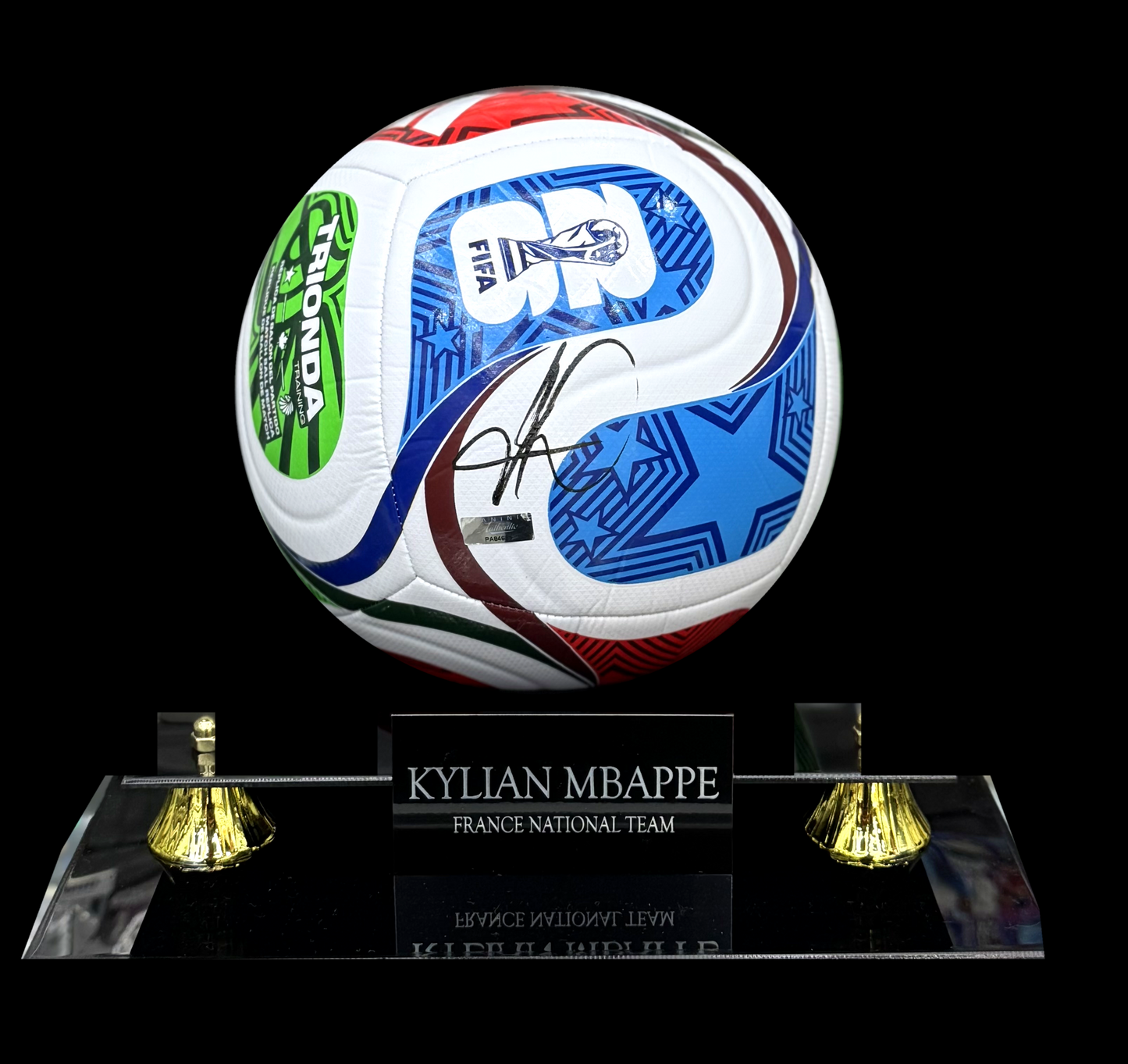 An autographed Kylian Mappre France national team soccer ball signed in black ink, displayed on a clear acrylic stand with gold-colored bell-shaped feet against a solid black background.