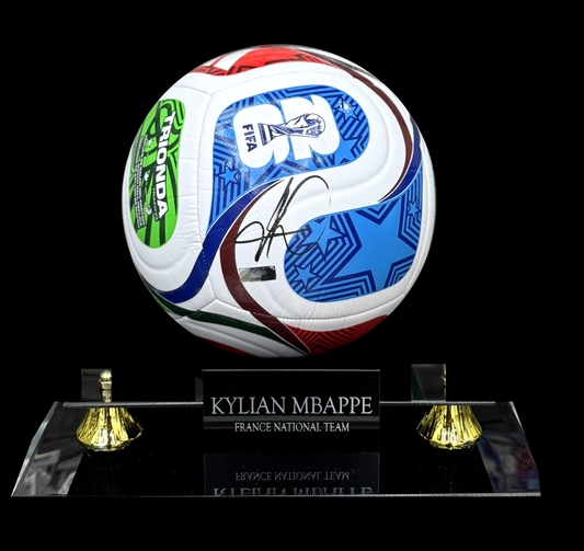 An autographed Kylian Mappre France national team soccer ball signed in black ink, displayed on a clear acrylic stand with gold-colored bell-shaped feet against a solid black background.