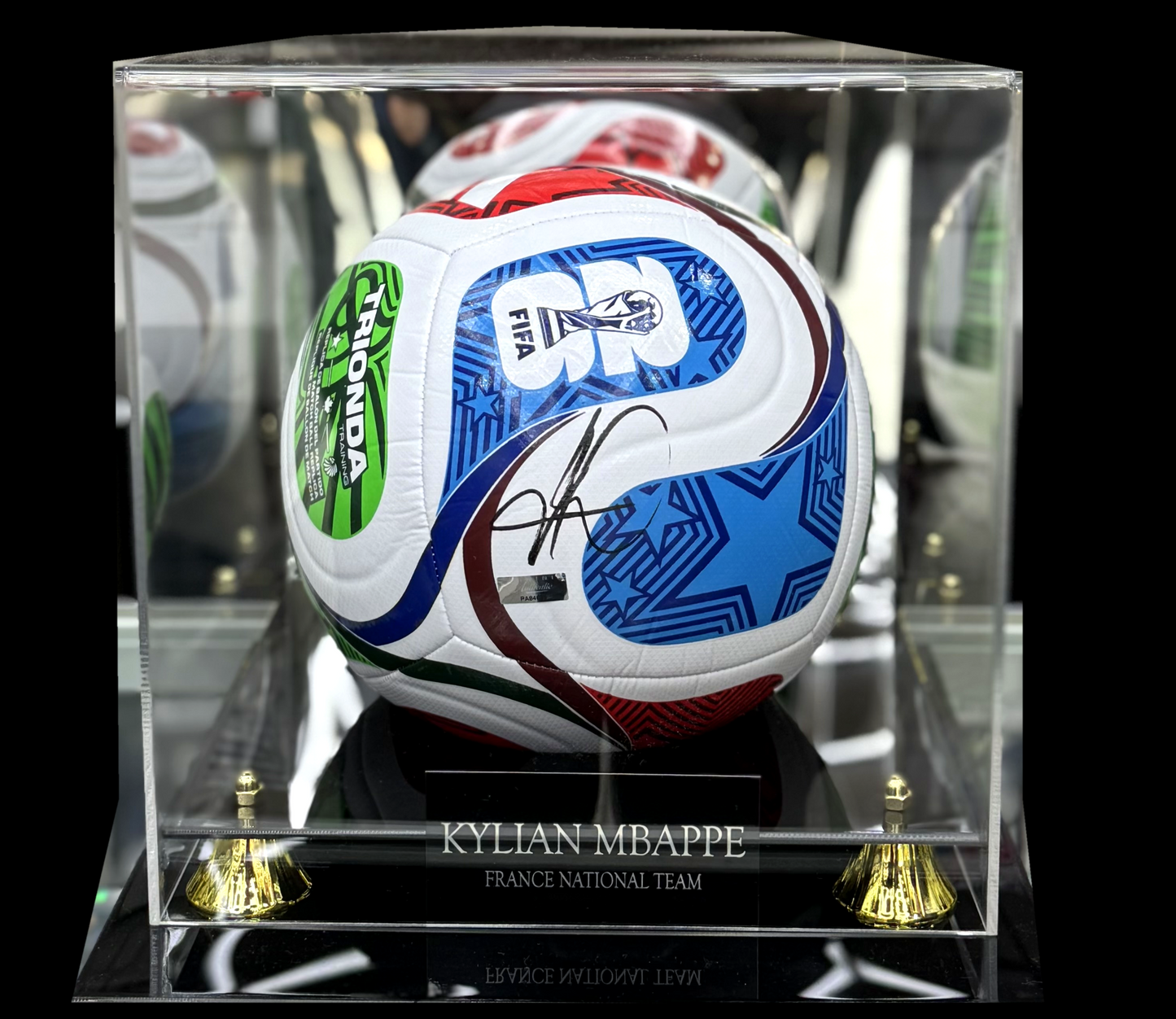 A signed soccer ball autograph display case featuring a white soccer ball with blue, red, and green graphics, signed in marker, housed inside a clear acrylic display box on a black background.